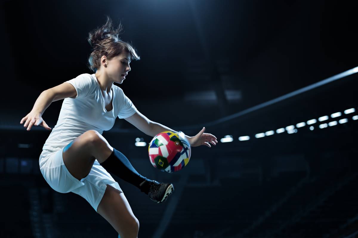 A player in white controls a football decorated with international flags in a floodlit stadium